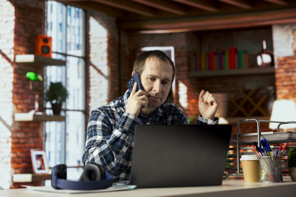 Stressed small business owner sitting at a desk in a home office, answering a phone call while working on a laptop, showing the challenges addressed through small business coaching, systems business coaching, small business coaching tools, and small business coaching framework